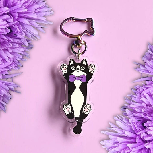 a tuxedo cat acrylic keyring with a moveable tail on the top of a purple background with purple flowers