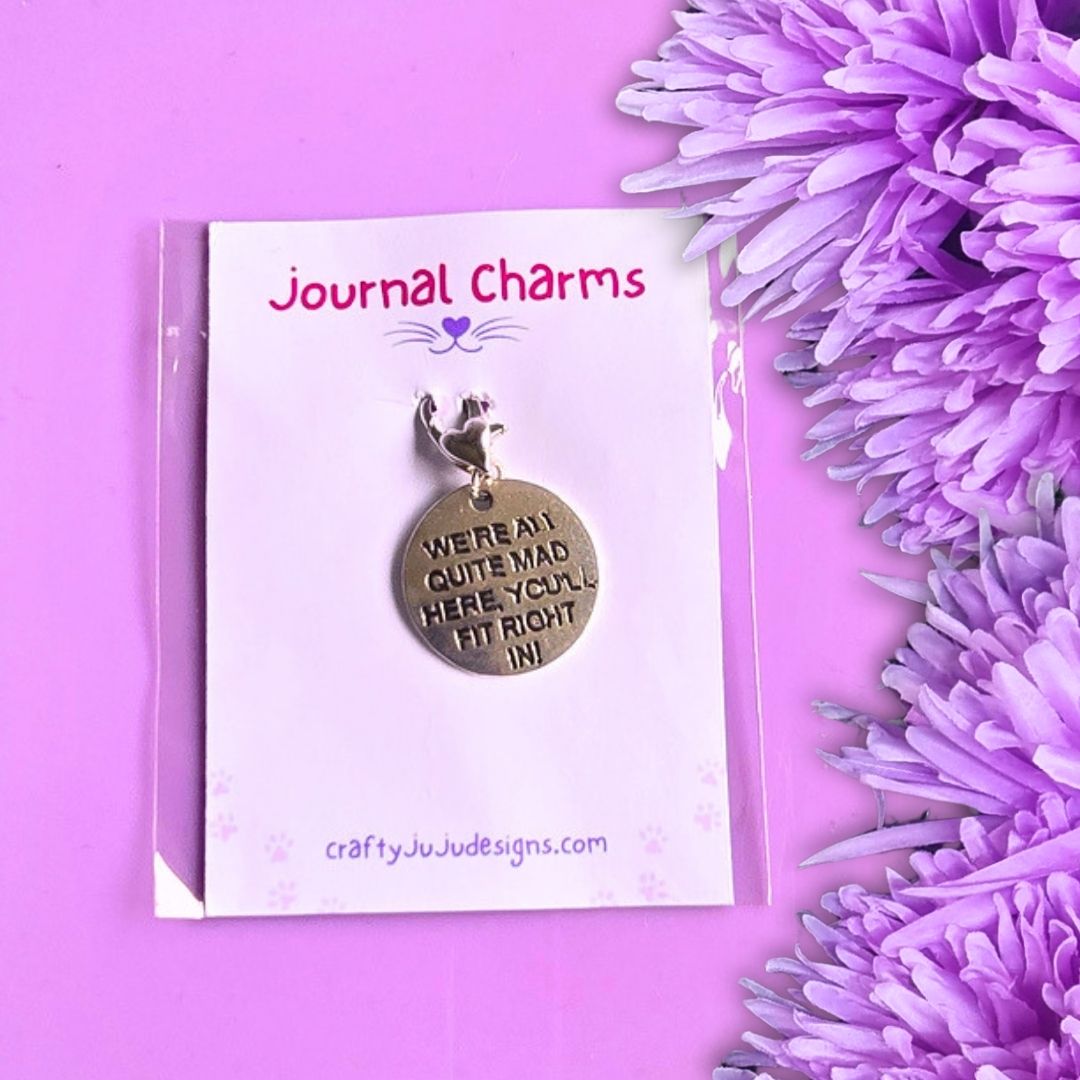 A round metal journal charm with engraved text reading 'WE'RE ALL QUITE MAD HERE YOU' FIT RIGHT IN' hangs from a small cat silhouette on a white display card labeled 'Journal Charms'. The card is photographed on a pink background with purple flowers along the right side.