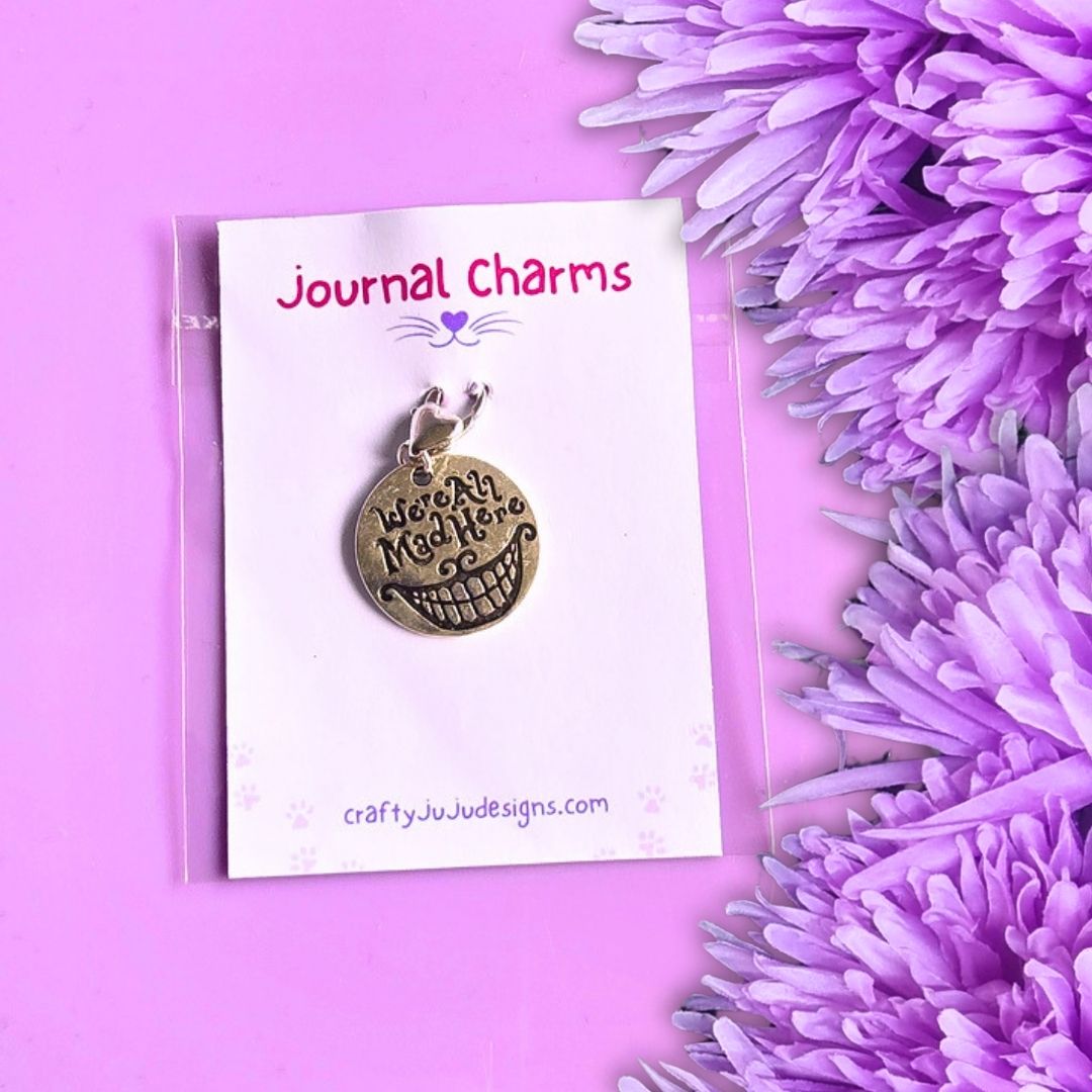 A round bronze-toned pendant charm with engraved text reading 'We're All Mad Here' and a smiley face, displayed on a white card labeled 'Journal Charms' against a pink background with purple flowers.