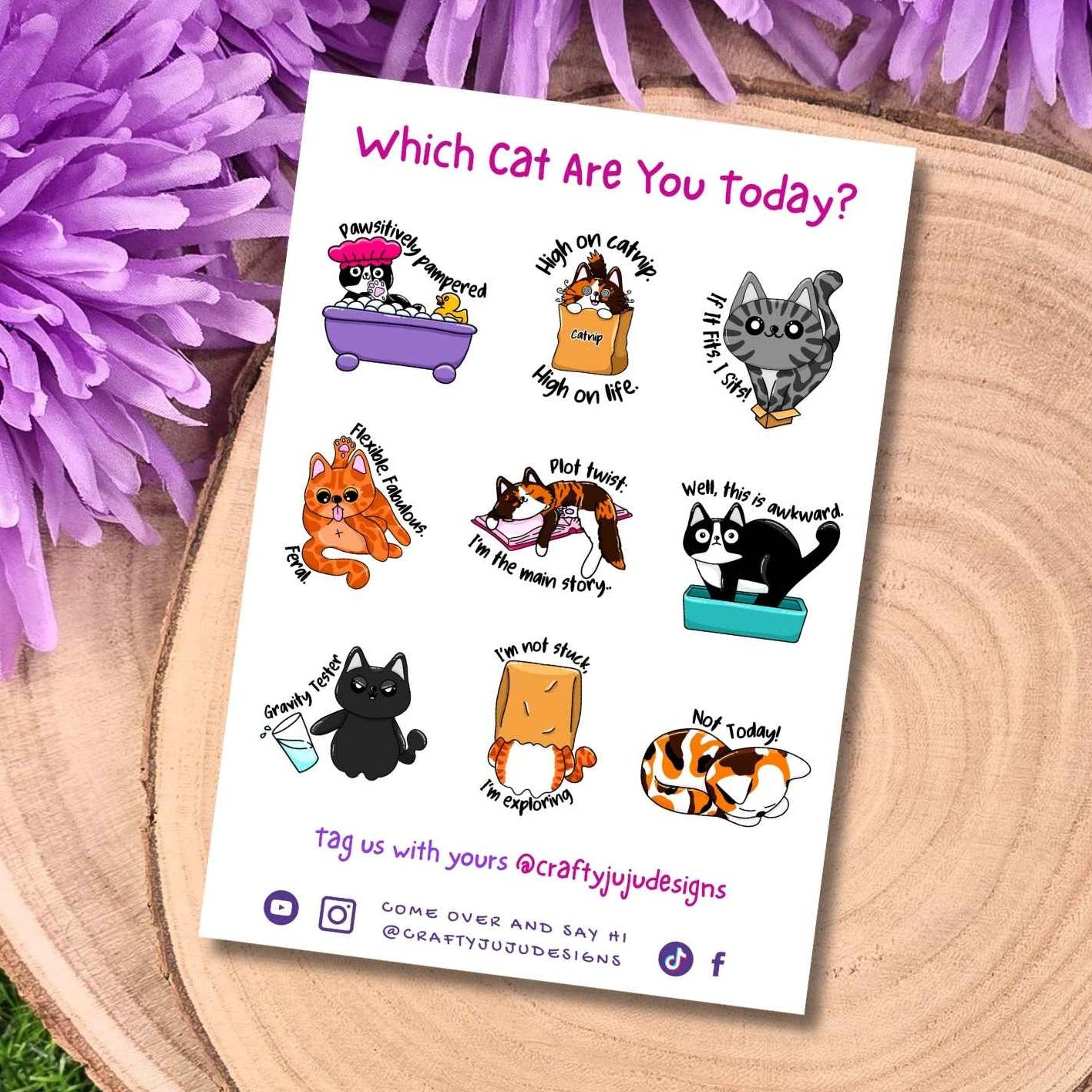 A printed sticker sheet titled "Which Cat Are You Today?" lies on a round wood slice surrounded by purple flowers and green grass. The sheet contains multiple small cartoon cat illustrations with humorous captions and social media handles.